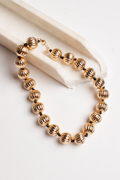 A gold-toned beaded necklace with ribbed, textured spherical beads and small dark spacer beads, displayed on a cream fabric prop against a white background.