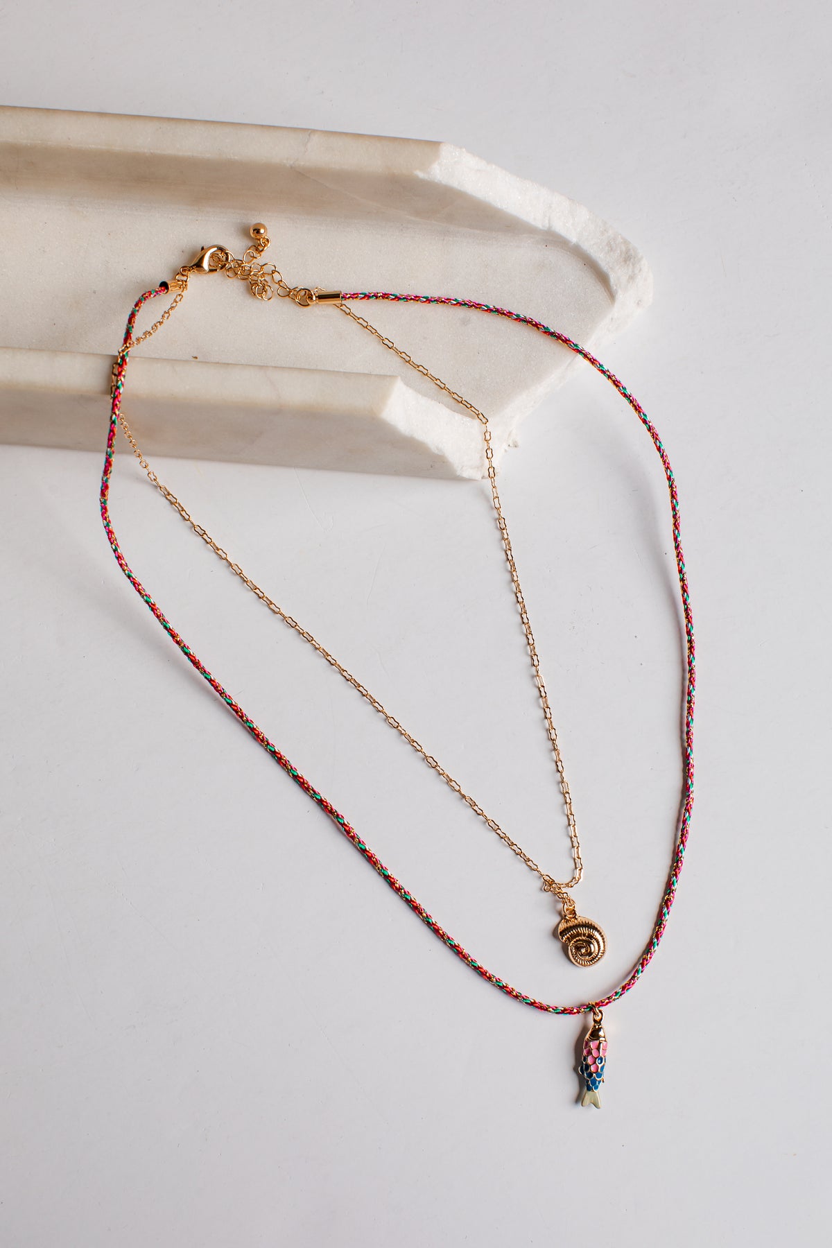 A delicate layered necklace set featuring a gold chain with a small gold spiral pendant and a longer red-and-gold beaded chain with a small multicolor fish charm, displayed on a light gray surface next to a white ceramic tray.
