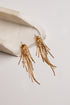 A pair of gold-toned dangling earrings with multiple thin fringe strands and small clear rhinestone clusters, displayed on a light neutral surface beside a curved cream stone prop.