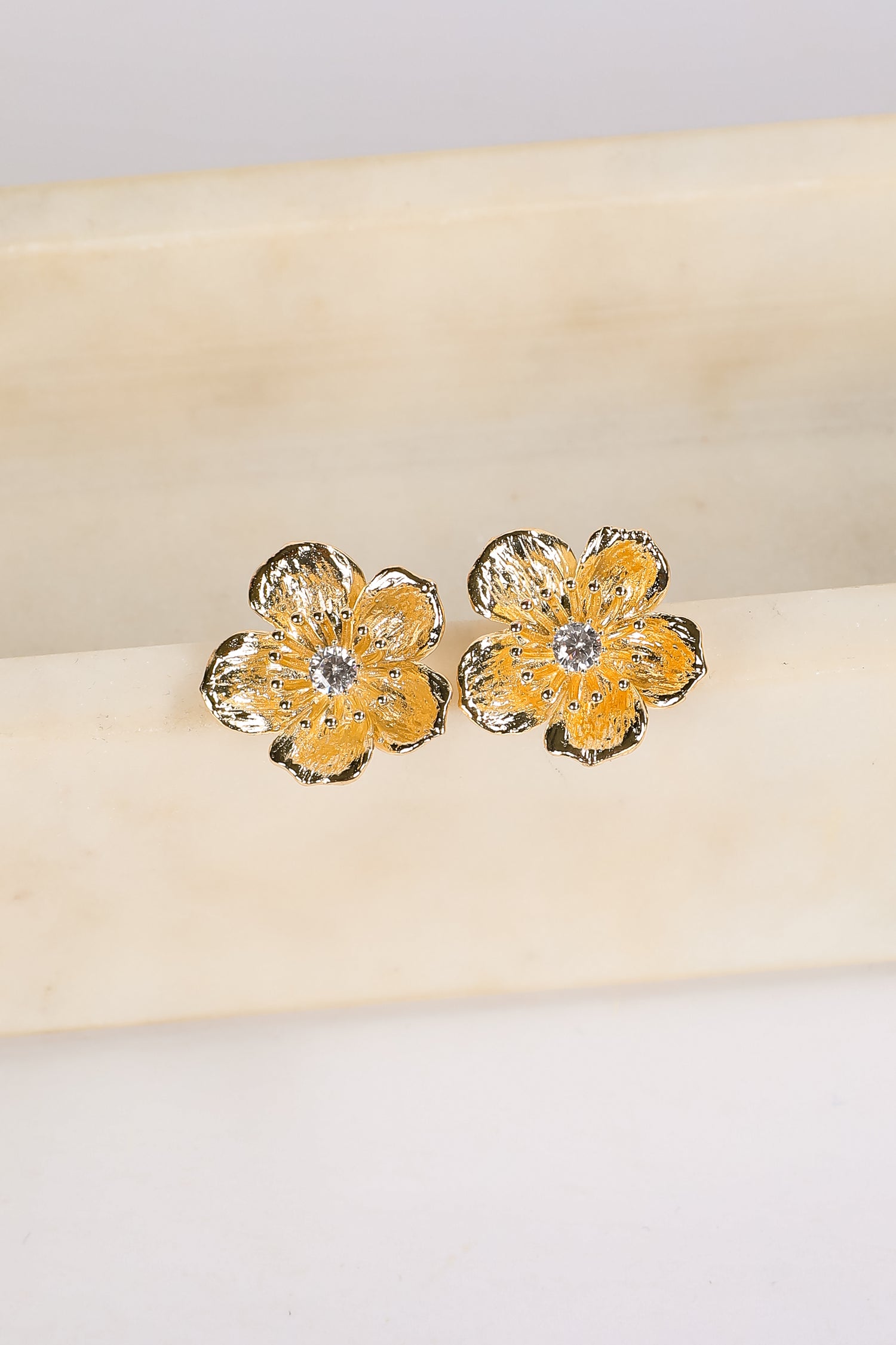 Gold flower-shaped earrings with a central gemstone on a beige cushion.