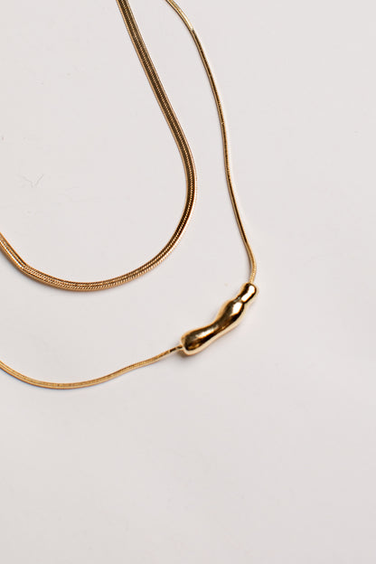 A gold snake chain necklace with a small, polished gold bead pendant, photographed on a clean, light off-white background.