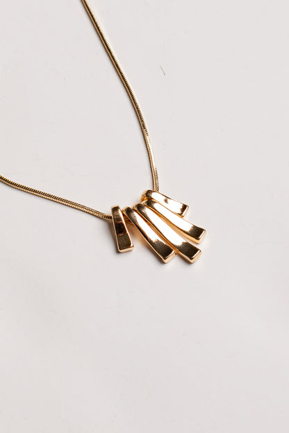 A gold necklace with a modern pendant featuring four overlapping rectangular bars, photographed on a clean, light off-white background.