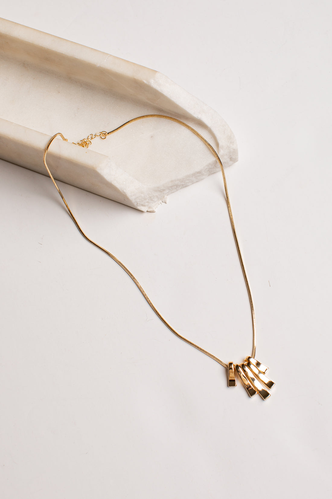 A delicate gold chain necklace with a small cluster of geometric bar pendants, styled on a light beige stone prop against a clean white background.