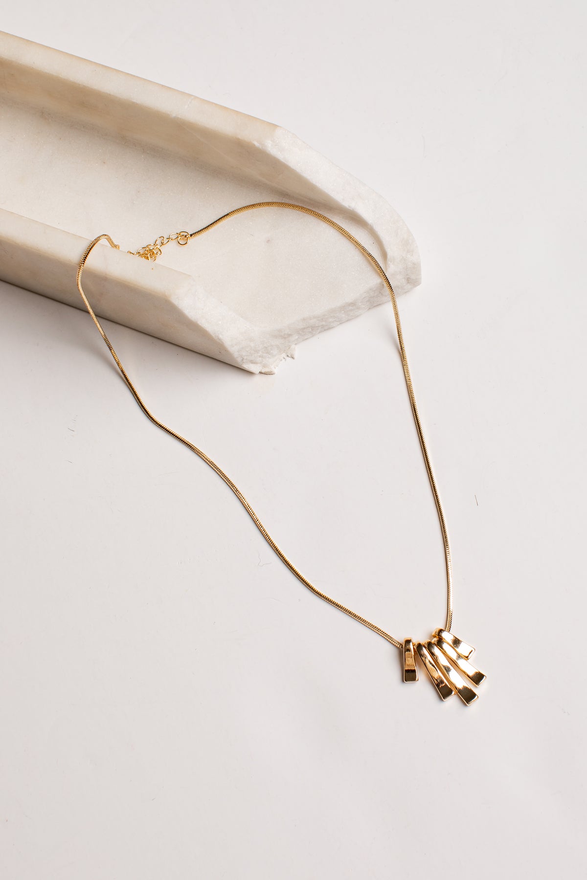 A delicate gold chain necklace with a small cluster of geometric bar pendants, styled on a light beige stone prop against a clean white background.
