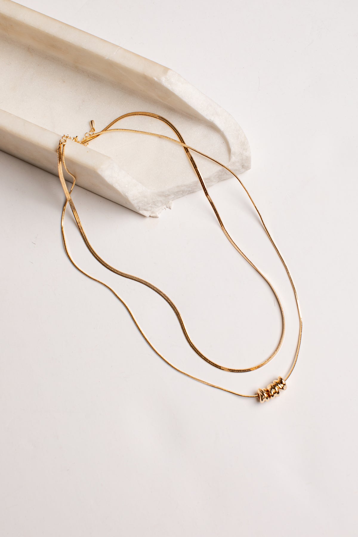 A delicate gold double-layer necklace with a small beaded accent on one layer, draped over a curved white stone prop on a light beige background.