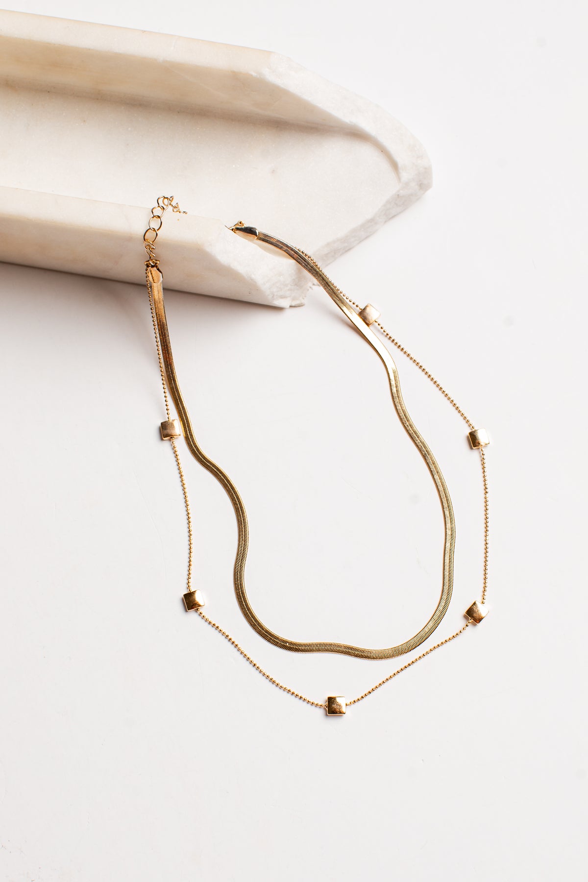 A double-layered gold necklace featuring a textured snake chain and a delicate chain with small gold heart charms, displayed on a light cream-colored wooden block against a white background.
