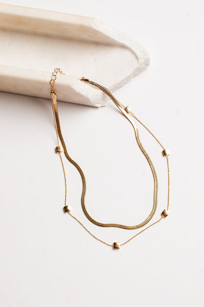 A double-layered gold necklace featuring a textured snake chain and a delicate chain with small gold heart charms, displayed on a light cream-colored wooden block against a white background.