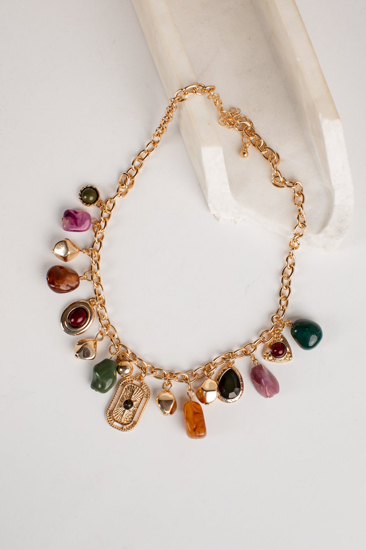 A gold-tone charm necklace with multiple colorful gemstone and crystal pendants, displayed on a white surface next to a cream-colored curved prop.