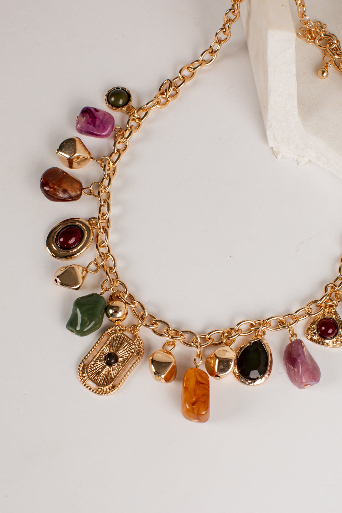 A gold-tone charm necklace featuring multiple colorful raw stone pendants in green, purple, brown, orange, black, and red, displayed on a clean white surface.