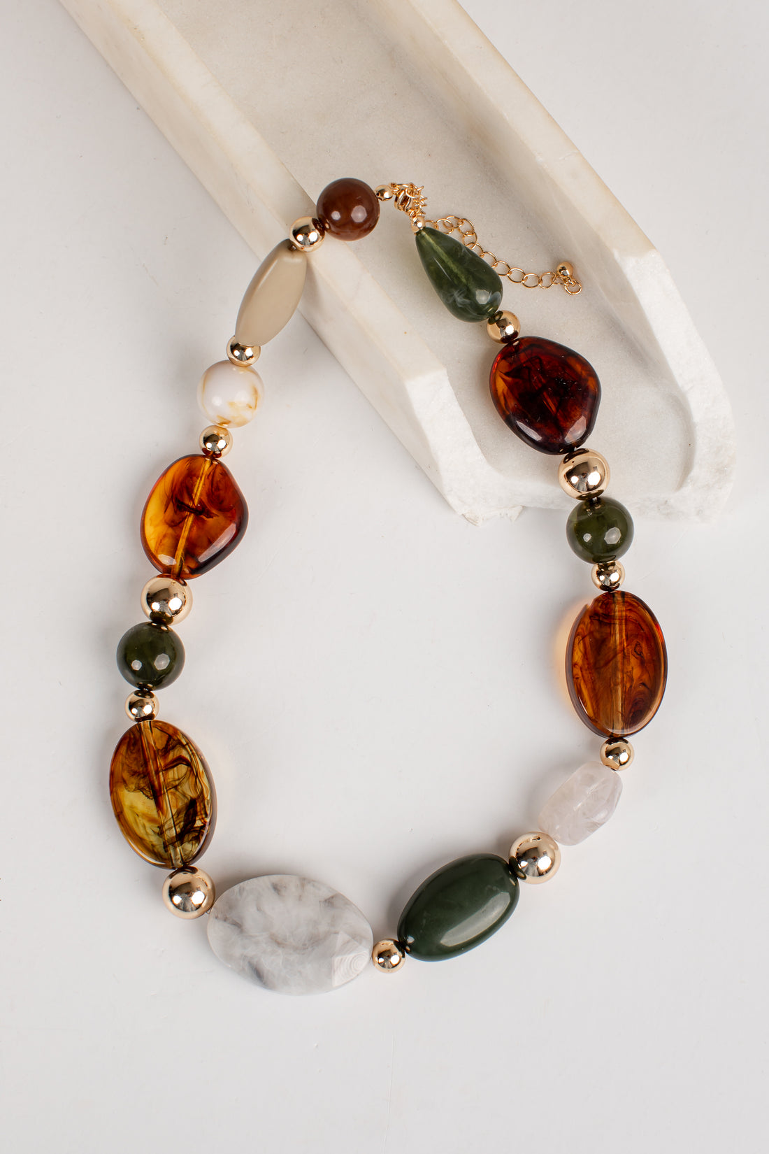 A beaded necklace with amber-brown, dark green, cream, and white stones, separated by small gold spacer beads, displayed on a white background with a cream-colored prop.