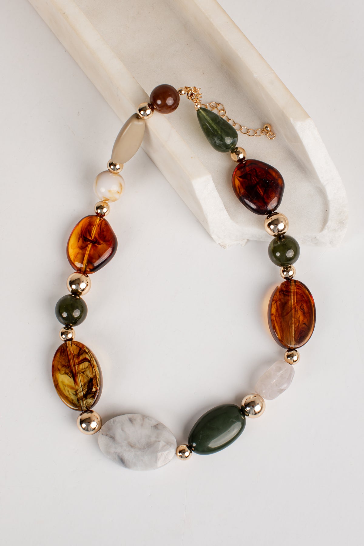A beaded necklace with amber-brown, dark green, cream, and white stones, separated by small gold spacer beads, displayed on a white background with a cream-colored prop.