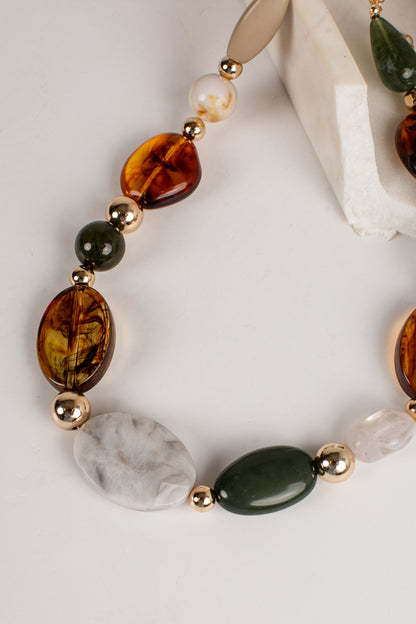 A close-up of a beaded necklace featuring large marbled amber-brown beads, a white pearl, a dark green stone, a white marbled stone, and polished gold spacer beads, arranged on a light neutral background.