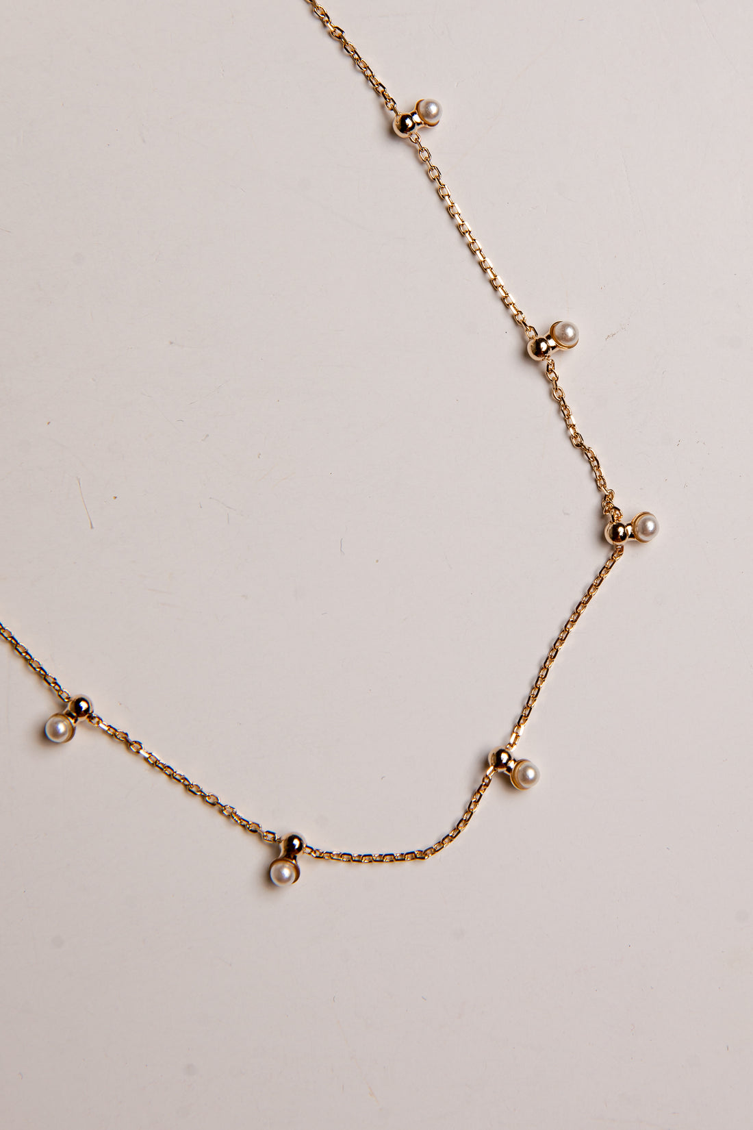 A delicate gold chain necklace featuring small pearl-like beads spaced along the chain, displayed on a plain off-white background.