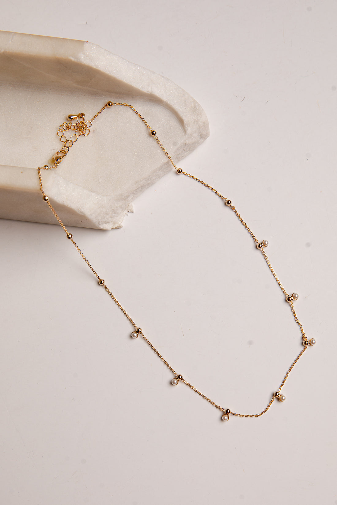 A delicate gold chain necklace with small bead accents and tiny clear stones, displayed on a light beige surface next to a textured cream stone prop.