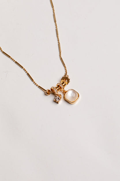 A gold chain necklace featuring a multi-charm pendant with a large square cream-colored stone, a small faceted gold bead, and a tiny clear crystal charm, displayed on a clean off-white background.