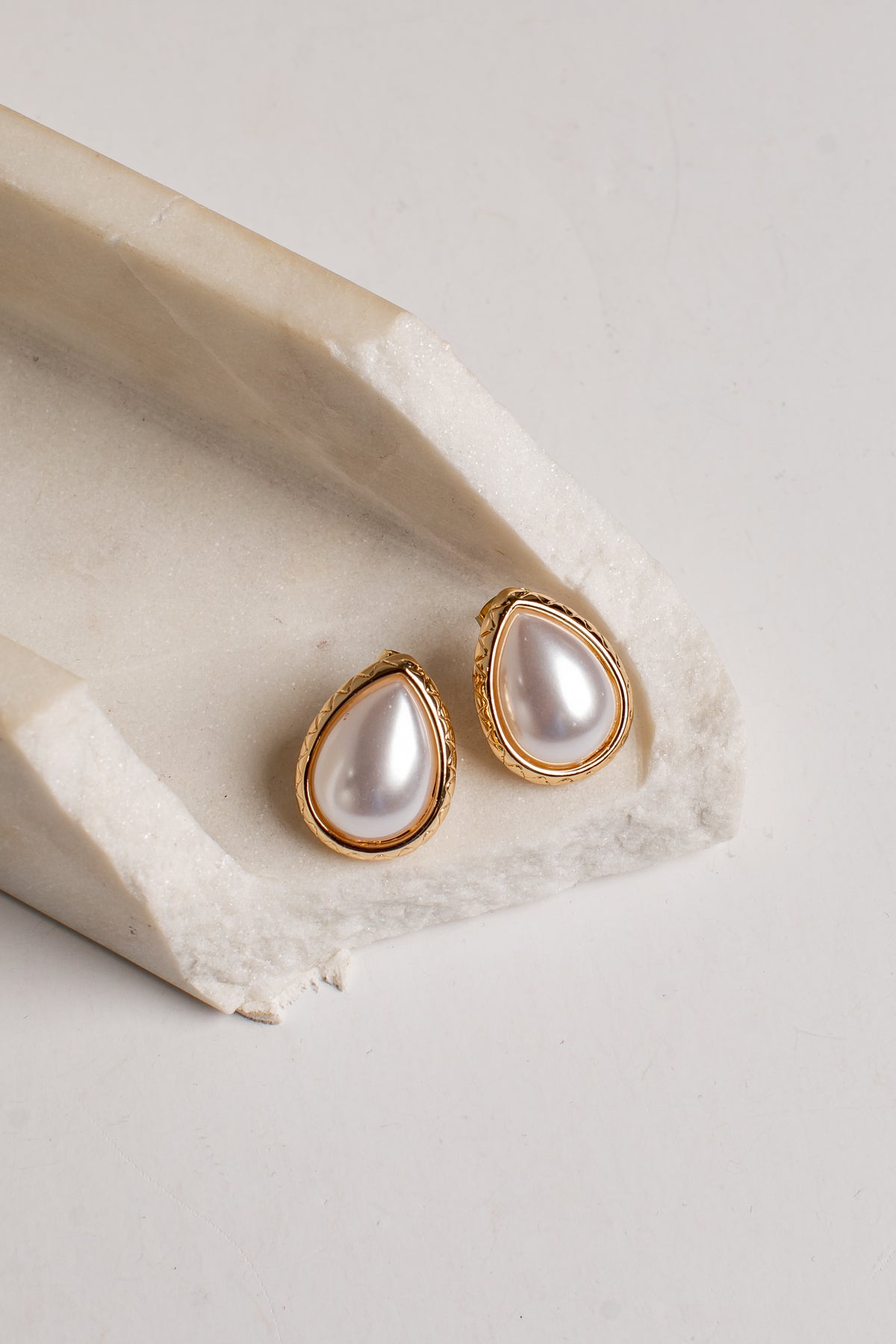 Teardrop-shaped gold stud earrings with white faux pearls, displayed on a textured beige stone riser against a light gray background.