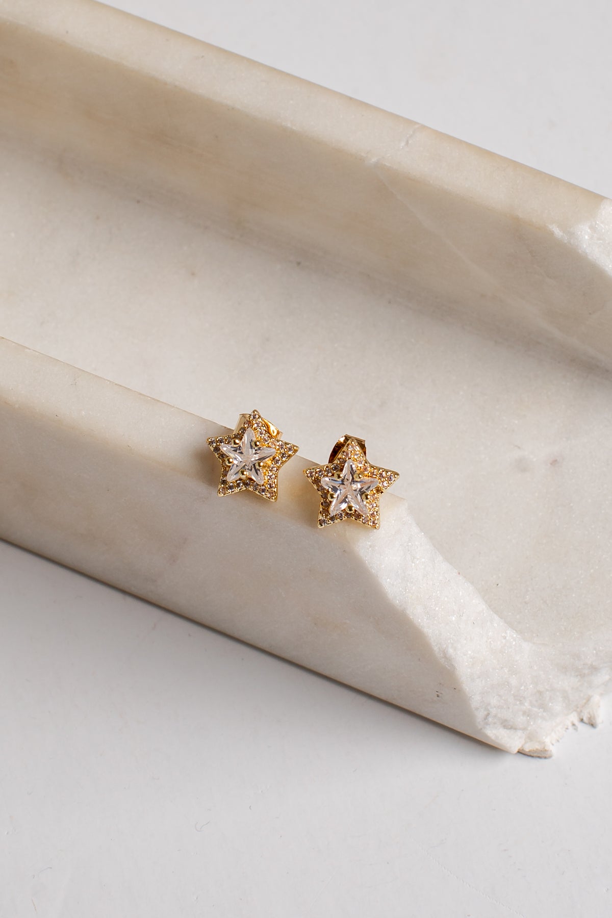 A pair of gold star-shaped stud earrings with clear gemstones, resting on a curved cream-colored stone slab against a light gray background.