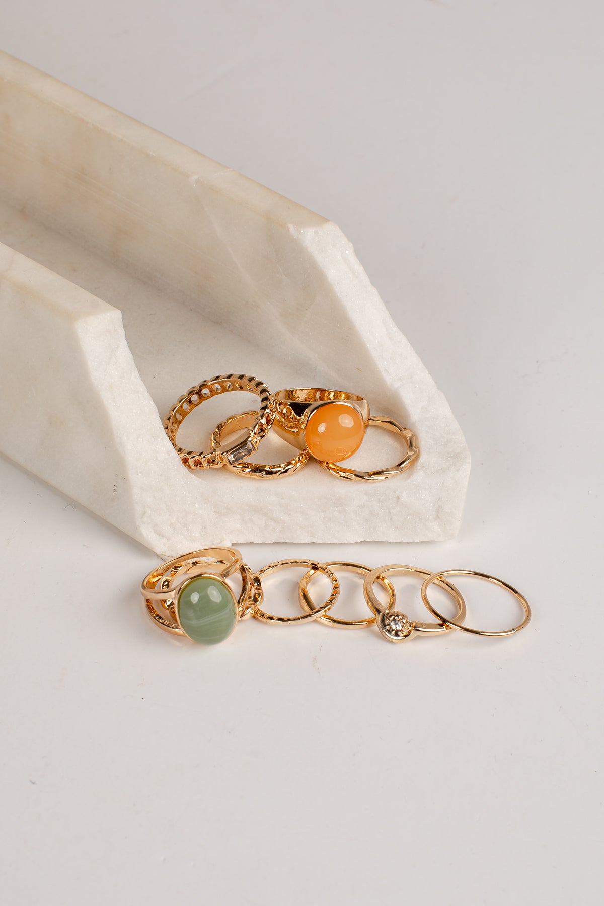 A set of gold rings displayed on a white marble ring stand and surface. The collection includes rings with textured bands, smooth orange and green stone bezels, and a delicate chain with a small clear crystal accent.