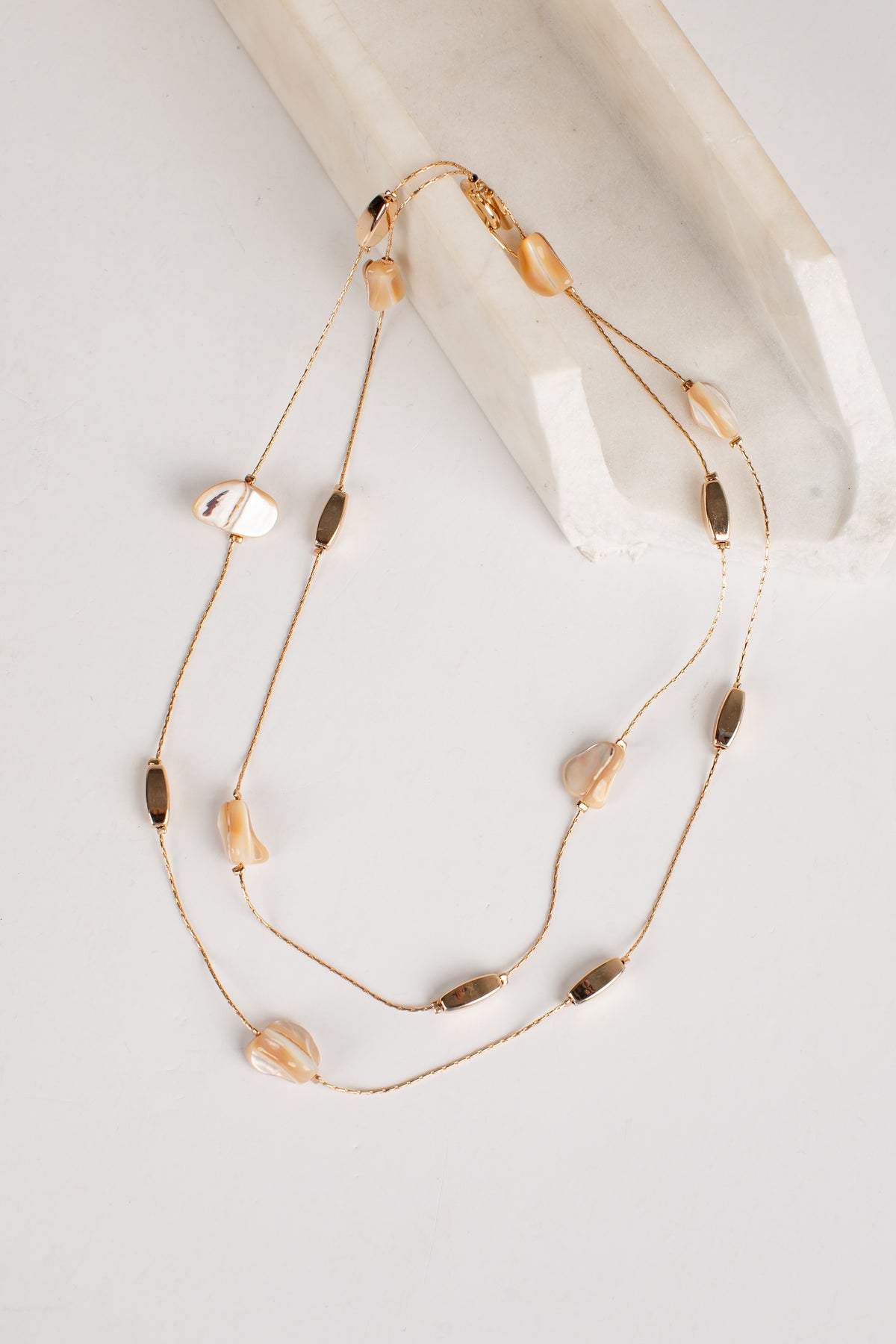 A double-strand gold-toned necklace with small metallic beads and irregular translucent shell-like accents, draped over a white sculptural prop on a light neutral background.