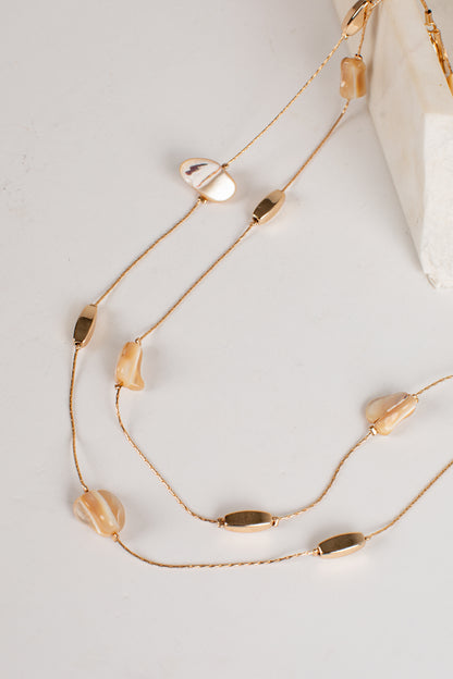 A gold-toned multi-strand necklace featuring irregular beige shell beads and polished oval bar accents, styled on a light gray surface with a white marble prop.