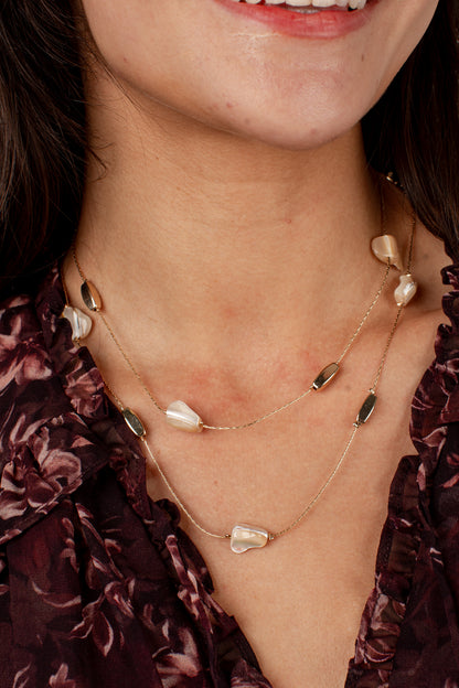 A close-up of a model wearing a double-layered gold necklace with irregular white pearl-like beads and small dark green marquise-cut beads, paired with a dark floral blouse.