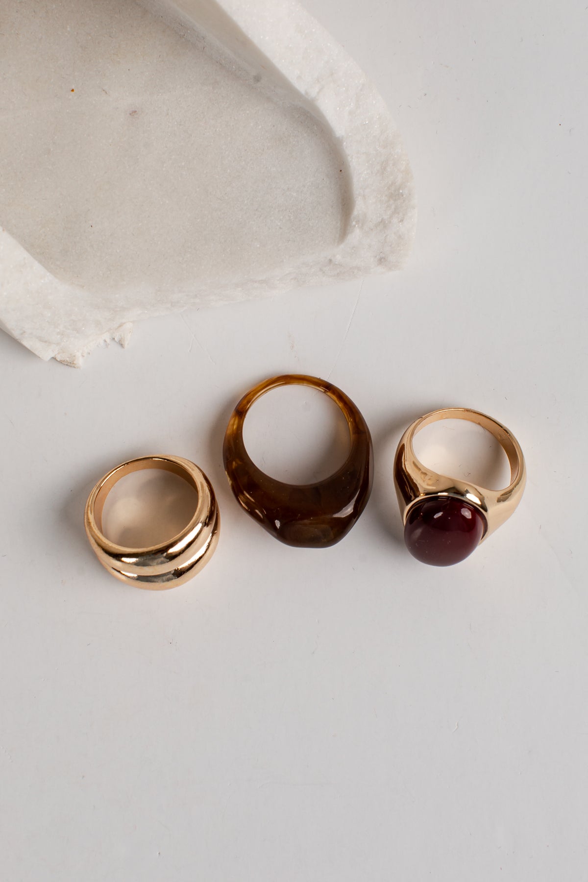 Three rings displayed on a light gray surface: a gold ring with three horizontal bands, a chunky translucent amber-brown ring, and a gold ring with a round dark red stone.