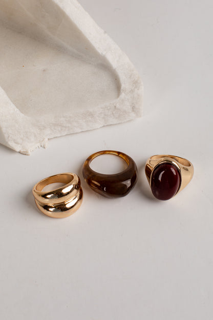 Three gold rings displayed on a light gray surface with a textured white stone prop. The rings include a double-band ring, a smooth dome-shaped ring, and a signet-style ring with an oval dark red stone.