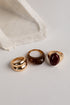 Three gold rings displayed on a light gray surface with a textured white stone prop. The rings include a double-band ring, a smooth dome-shaped ring, and a signet-style ring with an oval dark red stone.