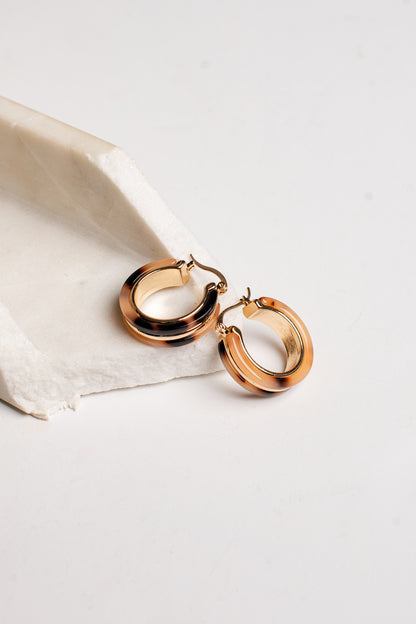 A pair of polished rose gold hoop earrings resting on a textured white stone slab against a clean white background.