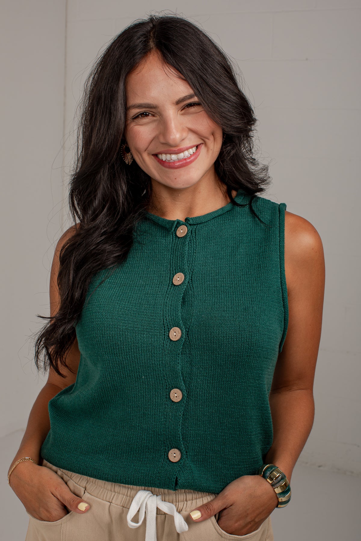 A smiling woman with long dark hair wearing a sleeveless dark green ribbed knit top with a button-down front, paired with beige drawstring pants, standing against a light gray wall.