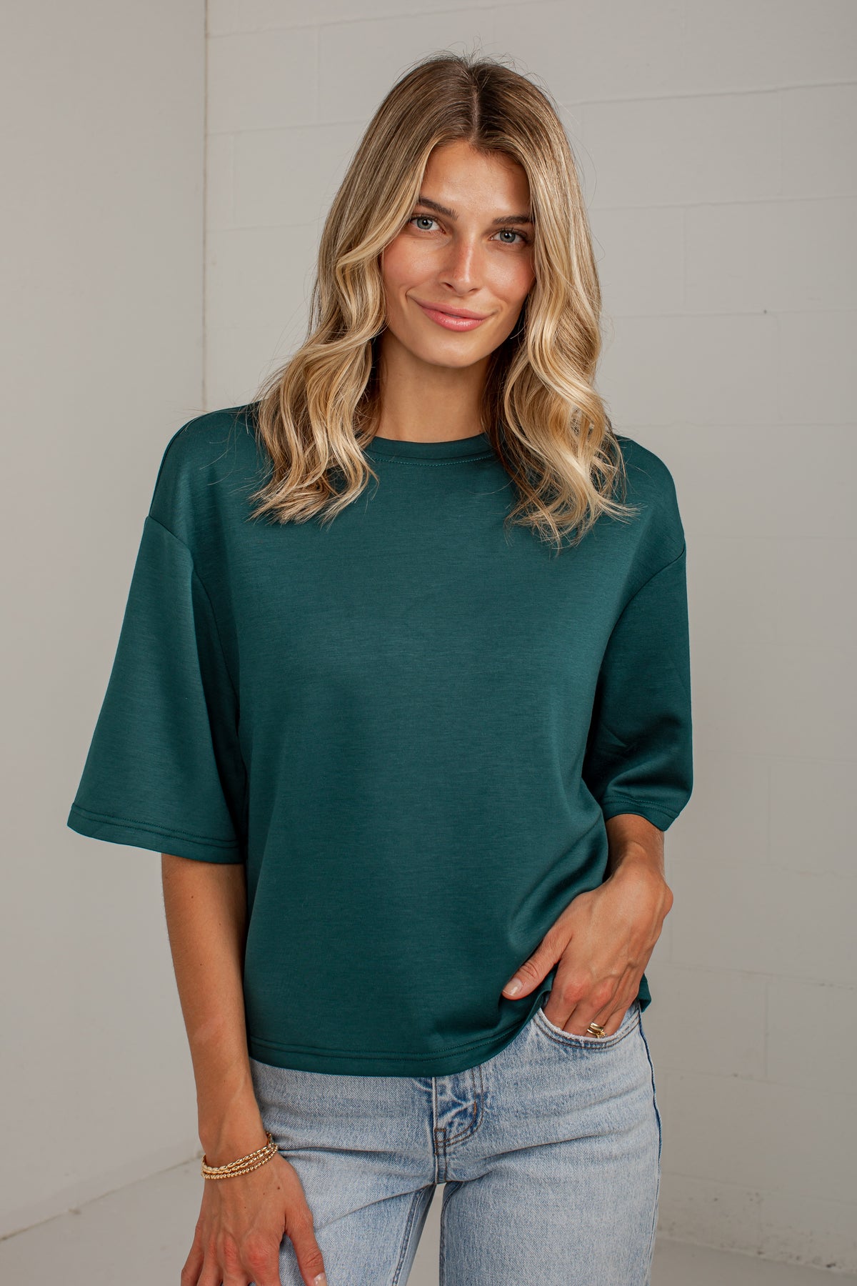 A woman with blonde wavy hair wearing an oversized dark teal green short-sleeve t-shirt and light wash blue jeans, standing against a white paneled wall.