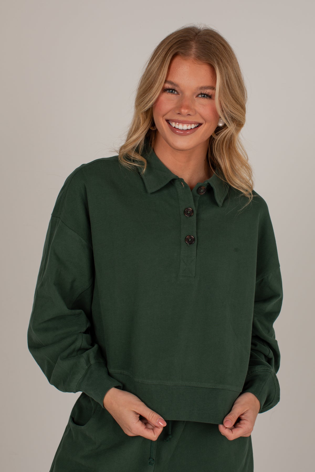 A smiling blonde woman wearing a dark forest green long-sleeve polo shirt with a buttoned collar, posed against a solid light gray studio background.