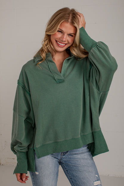 A woman wearing an oversized sage green half-placket pullover sweatshirt with balloon sleeves and distressed light-wash blue jeans, posing against a light gray wall.