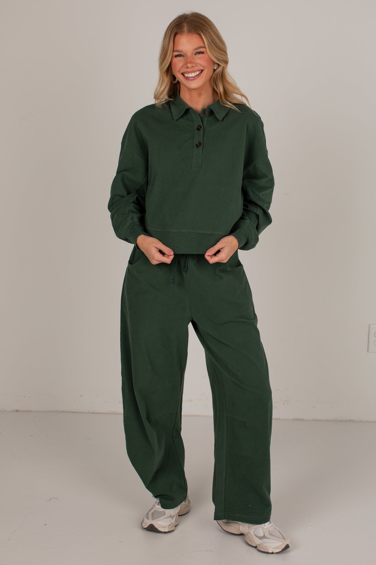 A woman modeling a dark green matching lounge set consisting of a long-sleeve button-up shirt and wide-leg pants, paired with white sneakers, against a plain light gray studio background.