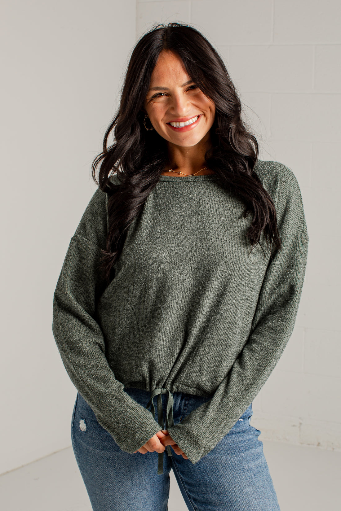 A woman modeling a loose, heathered sage green knit sweater with a tied hem, paired with blue jeans and brown heeled sandals, standing against a white studio wall.