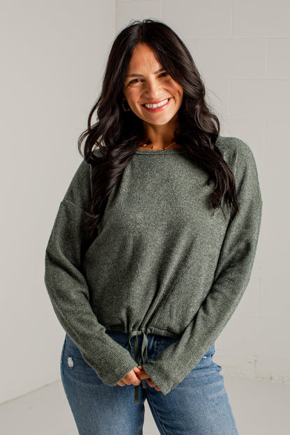 A woman modeling a loose, heathered sage green knit sweater with a tied hem, paired with blue jeans and brown heeled sandals, standing against a white studio wall.