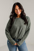 A woman modeling a loose, heathered sage green knit sweater with a tied hem, paired with blue jeans and brown heeled sandals, standing against a white studio wall.