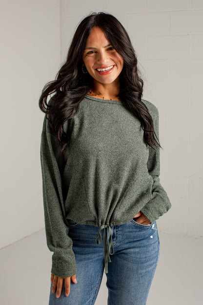 A woman modeling a loose, heathered sage green knit sweater with a tied hem, paired with blue jeans and brown heeled sandals, standing against a white studio wall.