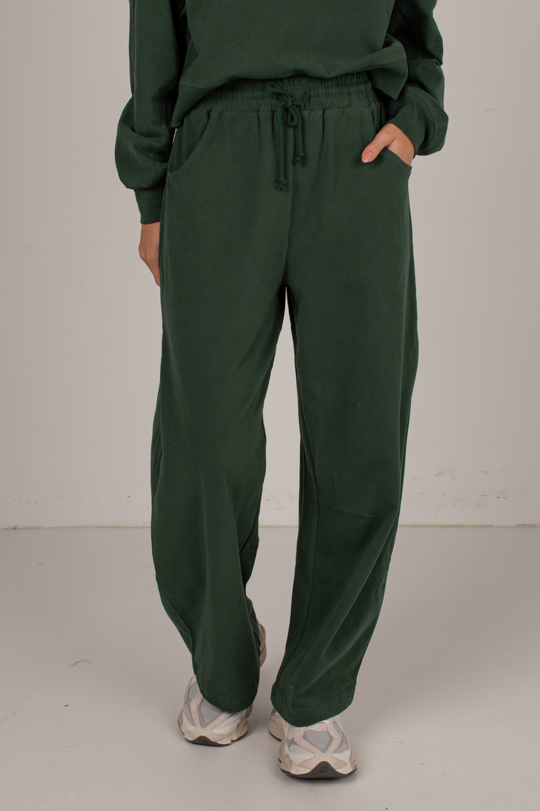 A person wearing dark green drawstring sweatpants with a matching sweatshirt, standing against a plain light gray studio background.