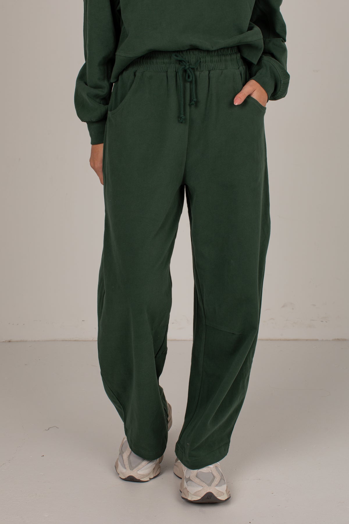 A person wearing dark green drawstring sweatpants with a matching sweatshirt, standing against a plain light gray studio background.