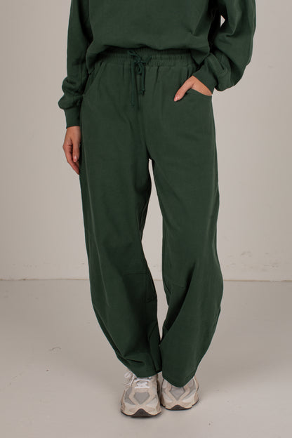 A person wearing dark green drawstring sweatpants with a matching sweatshirt and white sneakers, standing against a plain light gray studio background.