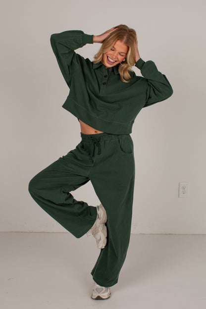 A woman modeling a dark green matching lounge set consisting of a cropped long-sleeve button-up shirt and wide-leg sweatpants, posing in a studio setting against a light gray wall.