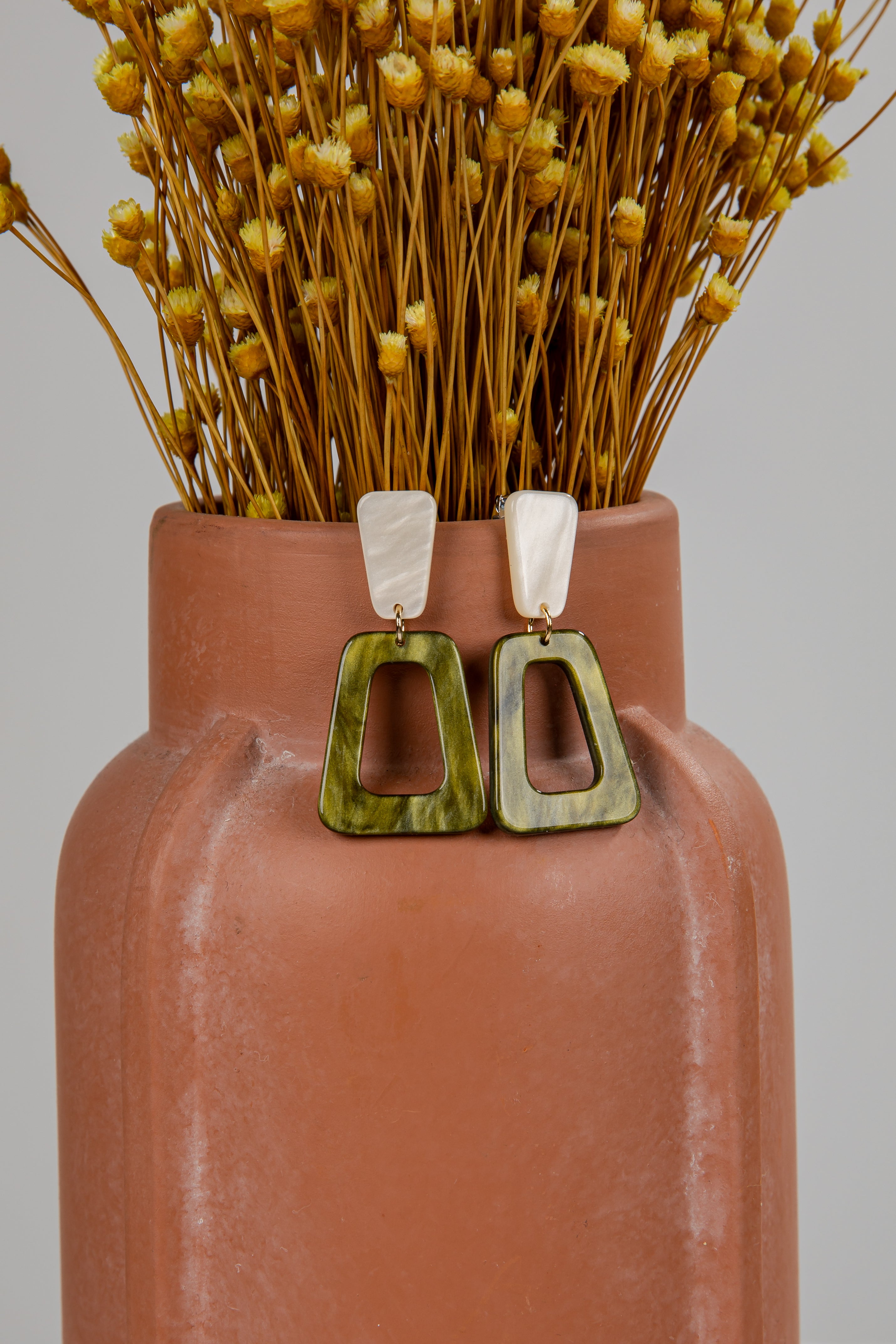 Terracotta vase with dried flowers and a pair of green earrings on a gray background