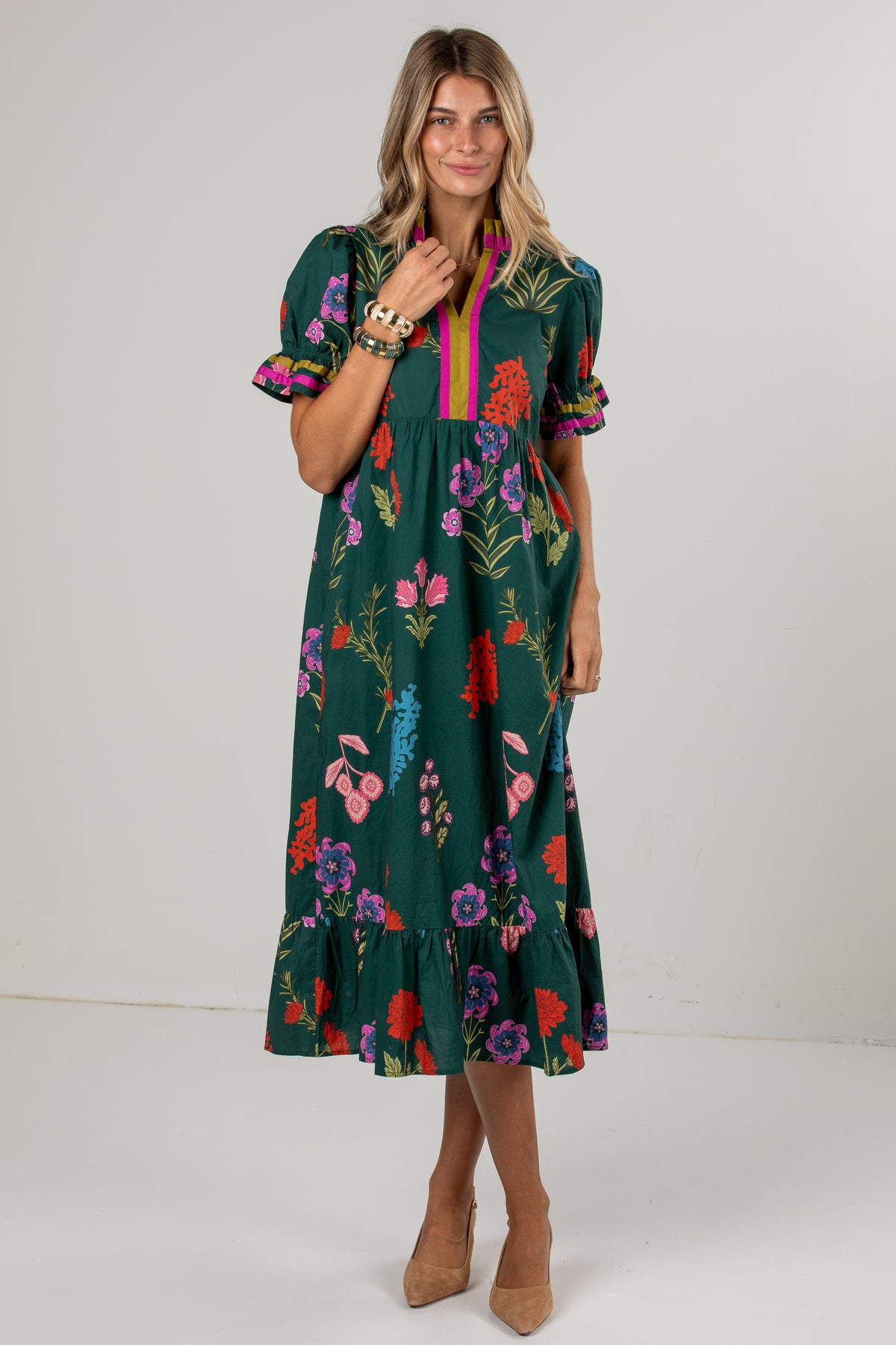 A woman modeling a dark green midi dress with a colorful floral print, puff sleeves, and a pink and yellow striped V-neckline, set against a plain light gray studio background.