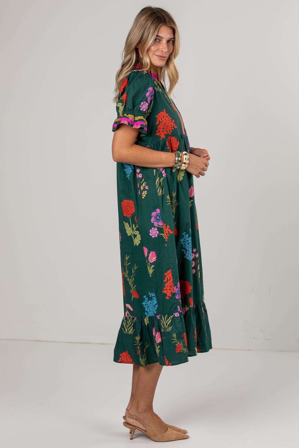A woman modeling a dark green midi dress with a colorful floral print, featuring short puff sleeves with ruffled trim and a tiered ruffle hem, paired with tan heels against a plain light gray studio background.