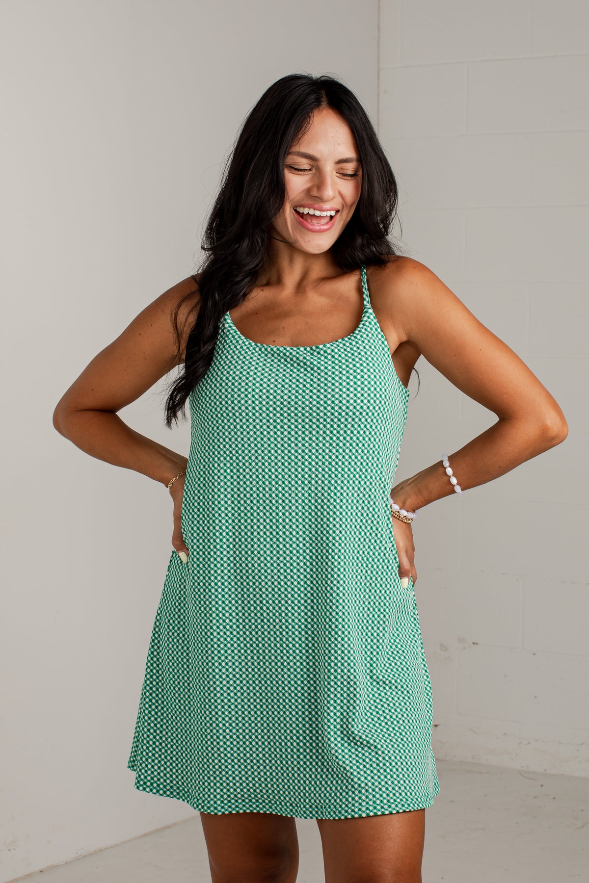 A woman modeling a sleeveless, scoop-neck mini dress with thin straps and side pockets, featuring a mint green and white geometric pattern, set against a plain light gray wall.
