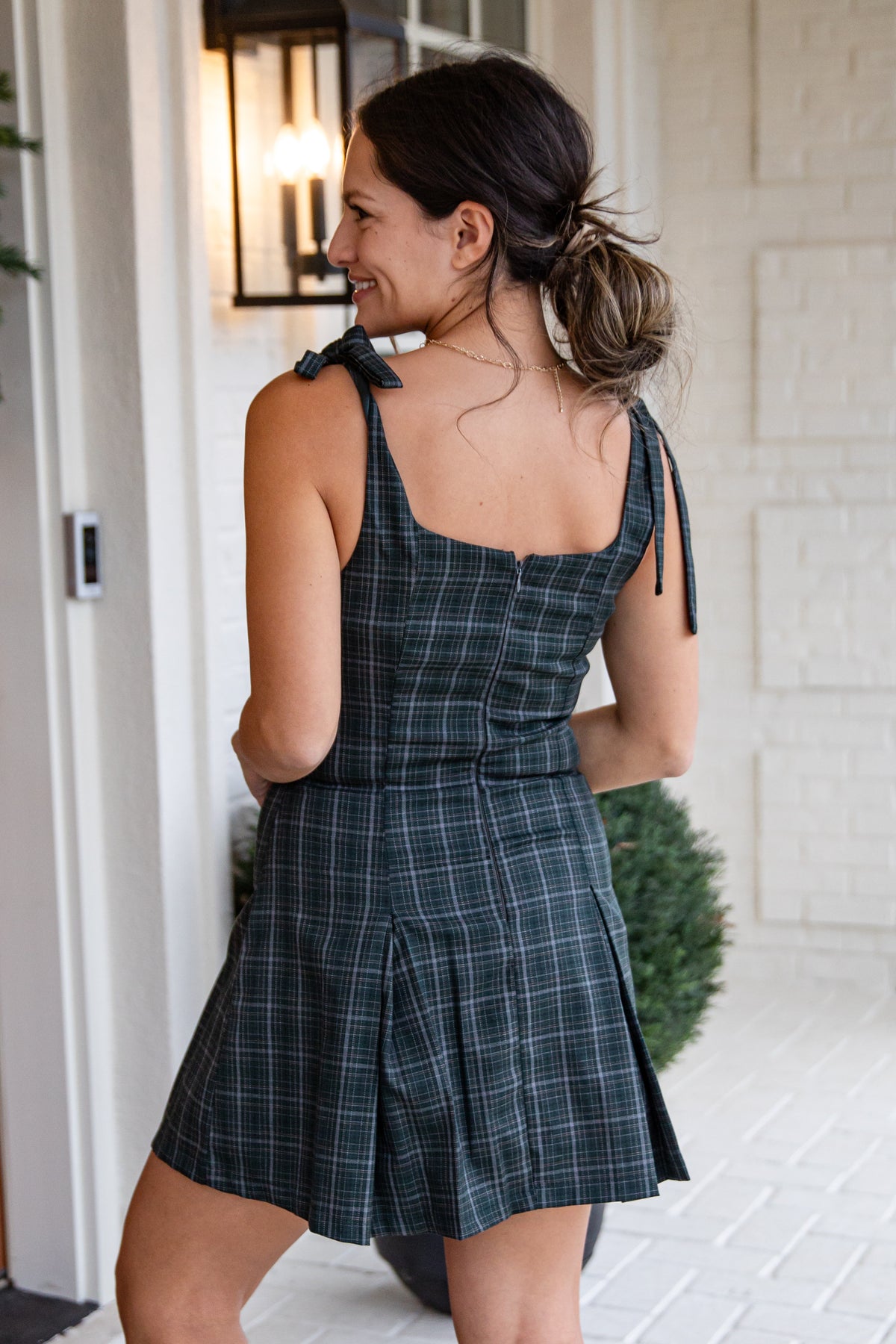 Woman wearing a plaid dress standing in a well-lit room with a white wall and light fixture.