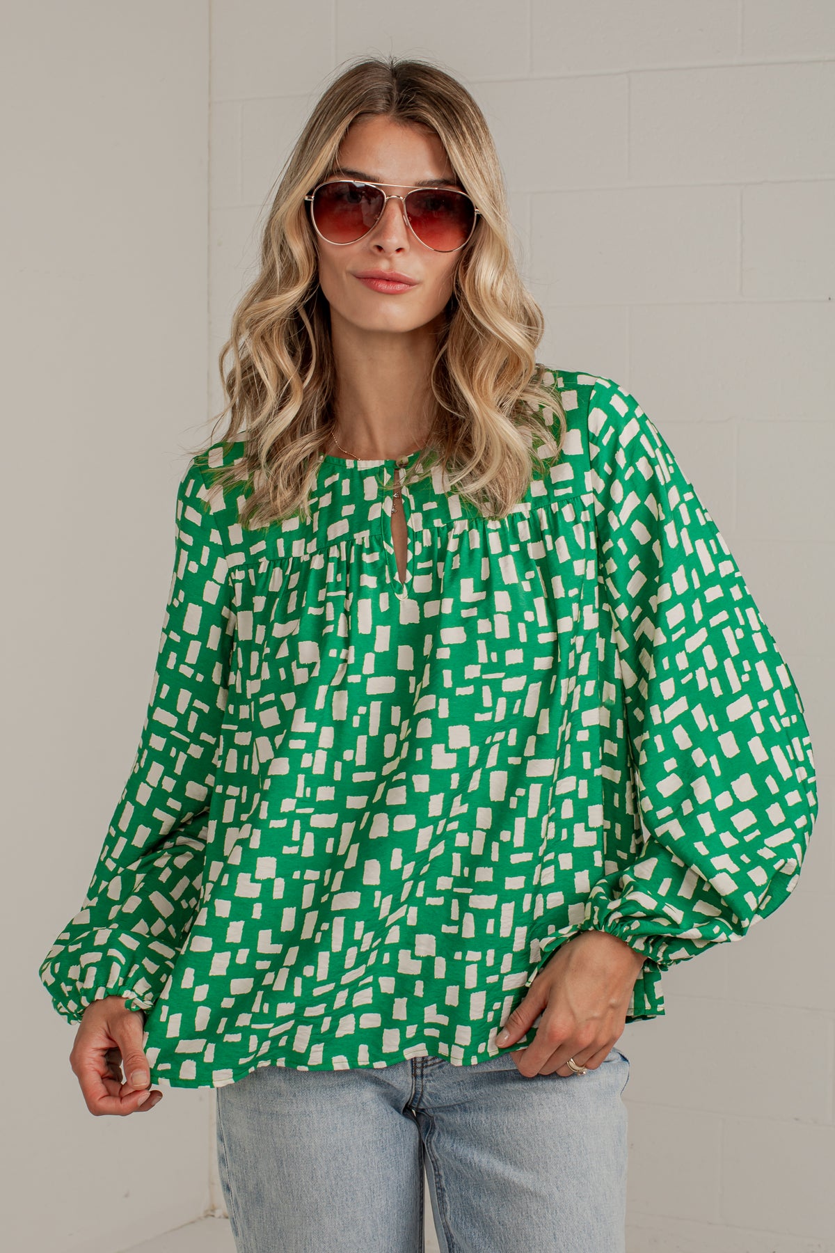A woman wearing a green and white geometric patterned blouse with balloon sleeves and a keyhole neckline, paired with light blue jeans and sunglasses, standing against a light gray brick wall.