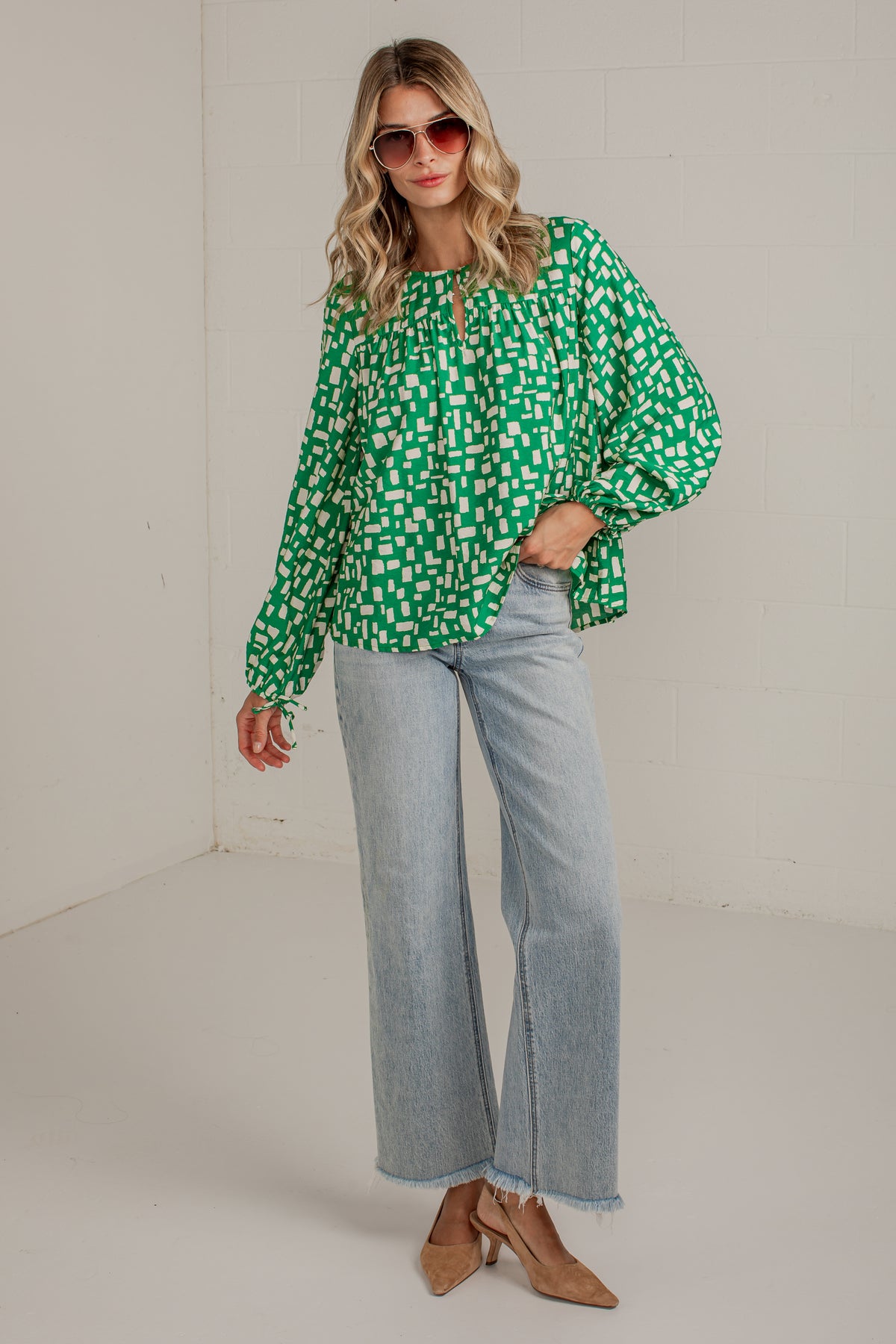 A woman modeling a green and white patterned long-sleeve blouse with light-wash wide-leg jeans and tan heeled sandals, standing in a plain indoor studio setting.