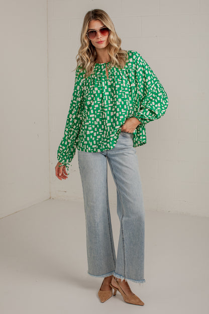 A woman modeling a green and white patterned long-sleeve blouse with light-wash wide-leg jeans and tan heeled sandals, standing in a plain indoor studio setting.
